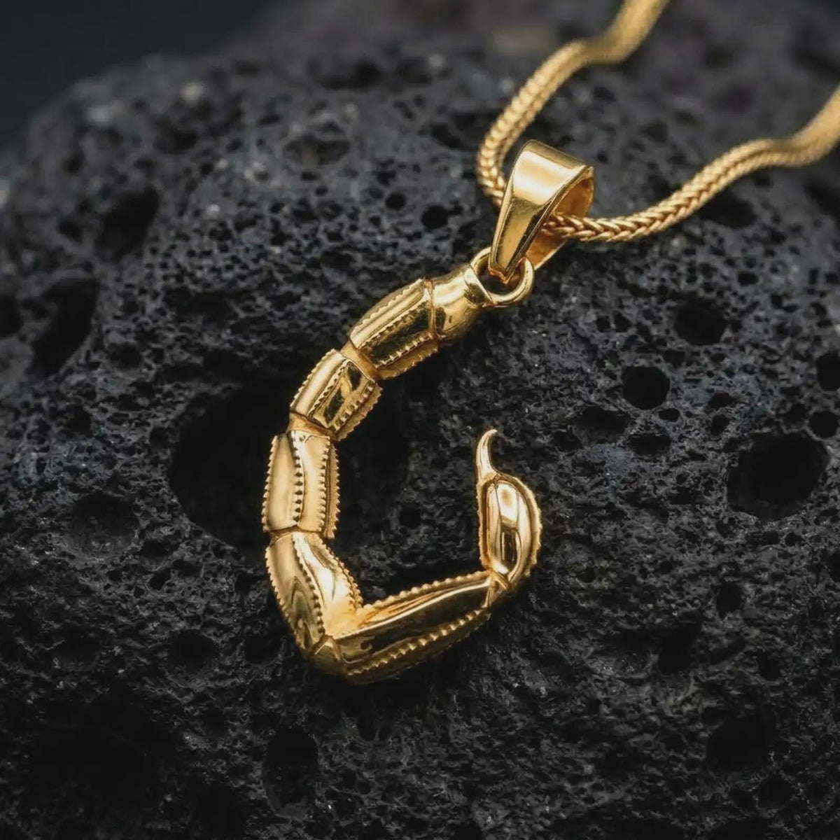 A gold plated scorpion tail pendant rests on rugged volcanic rock, featuring articulated armor-like segments, a curved stinger, and bold Scorpio-inspired symbolic styling in a luxury macro studio close-up.