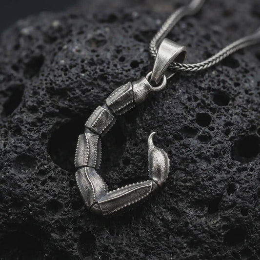 An oxidized sterling silver scorpion tail pendant rests on dark volcanic rock, showing articulated segmented sections, textured edges, a curved stinger, and dark recessed details in a dramatic macro studio close-up.