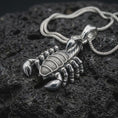 Load image into Gallery viewer, A polished sterling silver scorpion pendant is displayed on textured black volcanic rock, highlighting the dimensional claws, armored body segments, sculpted legs, and raised tail in a detailed macro photograph.
