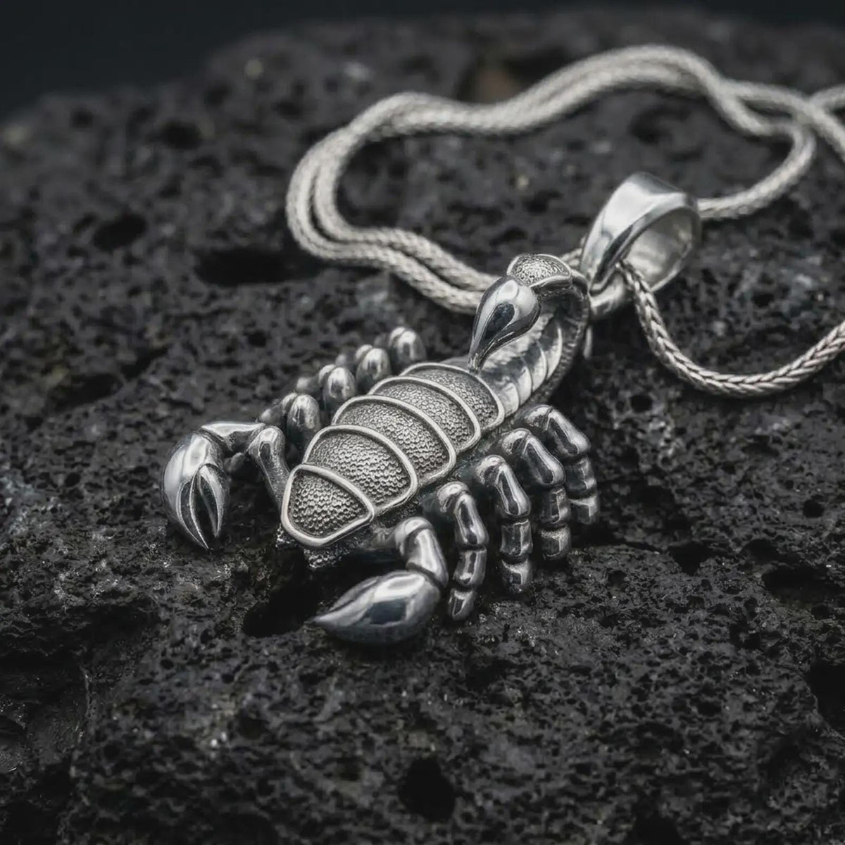 A polished sterling silver scorpion pendant is displayed on textured black volcanic rock, highlighting the dimensional claws, armored body segments, sculpted legs, and raised tail in a detailed macro photograph.
