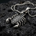Load image into Gallery viewer, An oxidized sterling silver scorpion pendant rests on dark volcanic rock, featuring detailed claws, segmented legs, textured body armor, curved tail, and pointed stinger in a dramatic macro studio close-up.
