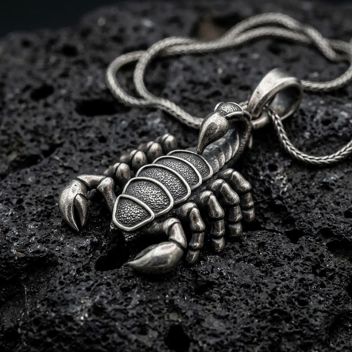 An oxidized sterling silver scorpion pendant rests on dark volcanic rock, featuring detailed claws, segmented legs, textured body armor, curved tail, and pointed stinger in a dramatic macro studio close-up.