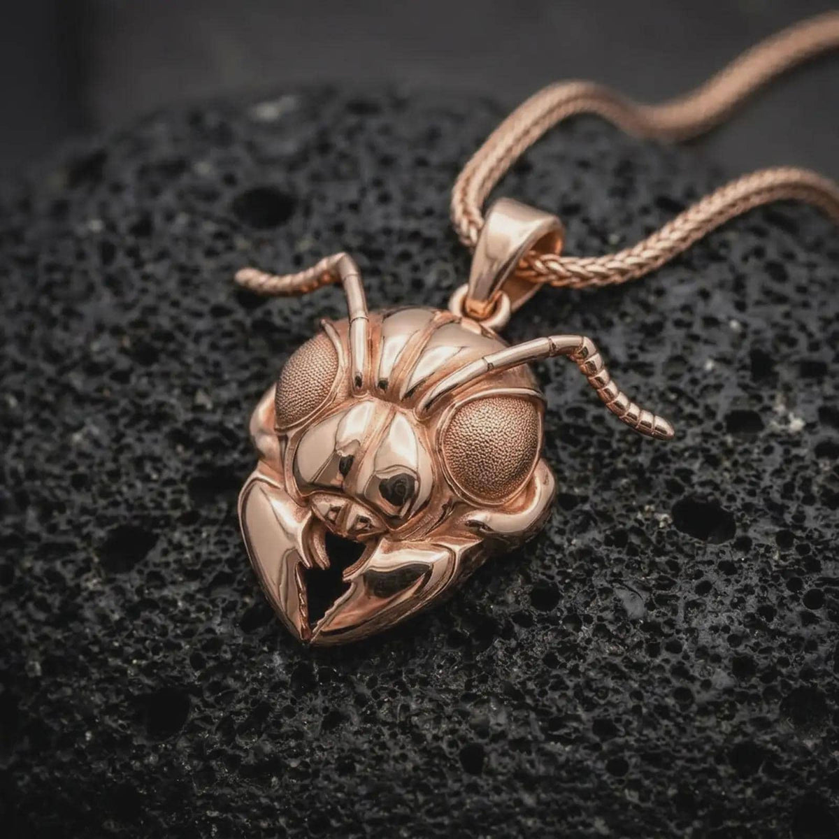 The rose gold plated insect necklace is arranged on black volcanic rock, emphasizing the ant-inspired head, textured eyes, curved antennae, dimensional facial structure, and polished statement pendant finish.