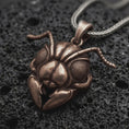Load image into Gallery viewer, A Bronze sterling silver ant head pendant rests on dark volcanic rock, featuring large textured compound eyes, segmented antennae, sculpted facial armor, and prominent mandibles in a dramatic macro close-up.
