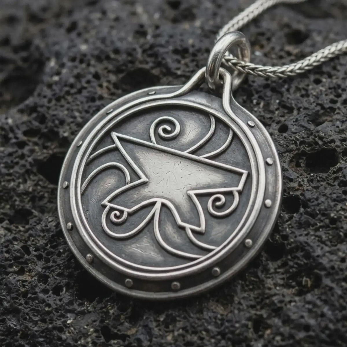 An oxidized sterling silver Zenithar-inspired amulet pendant rests on dark volcanic rock, featuring a raised geometric merchant symbol, spiral details, dotted border, and dark recessed circular background.