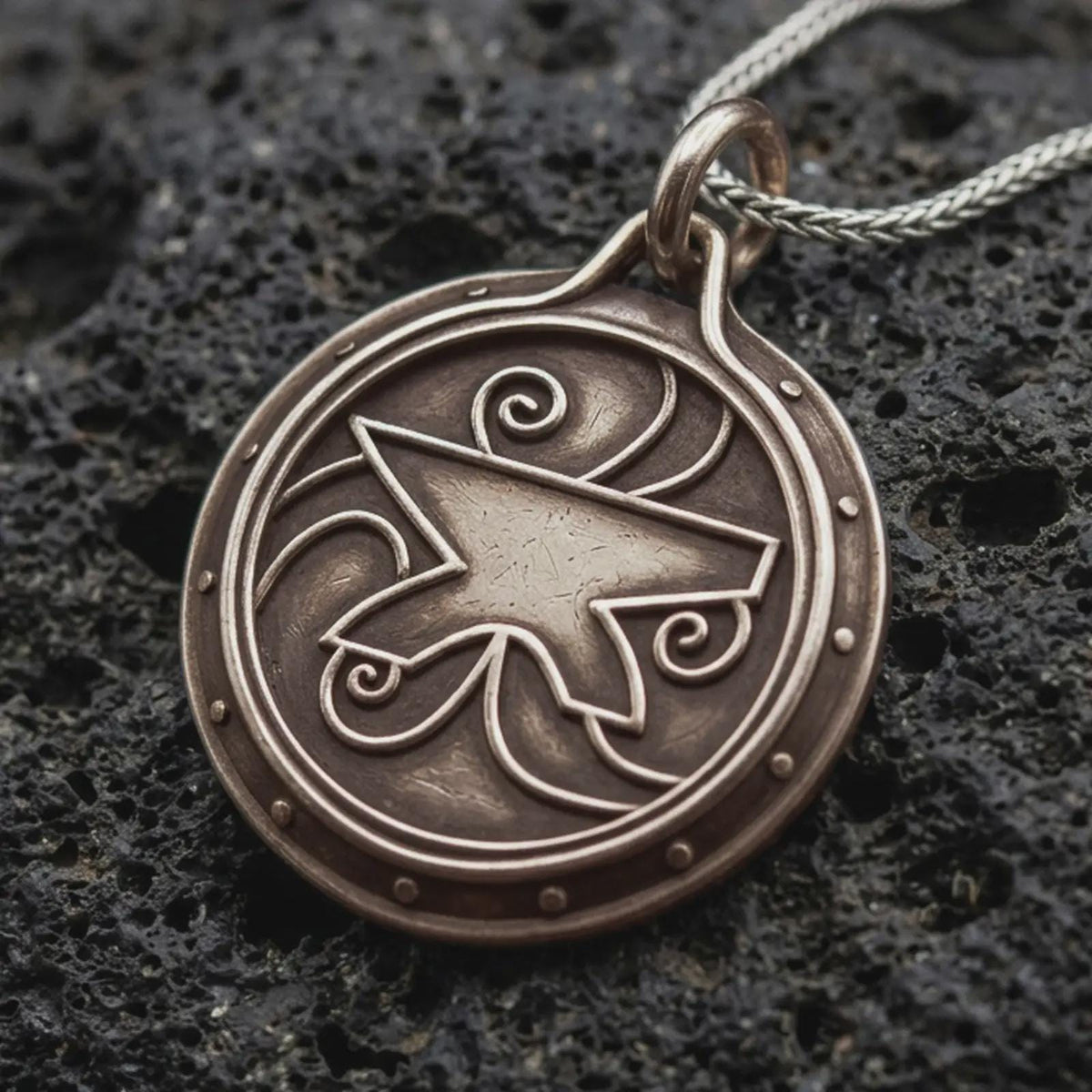 A bronze sterling silver Zenithar-inspired amulet pendant rests on dark volcanic rock, featuring a raised geometric merchant symbol, spiral details, dotted border, and dark recessed circular background.