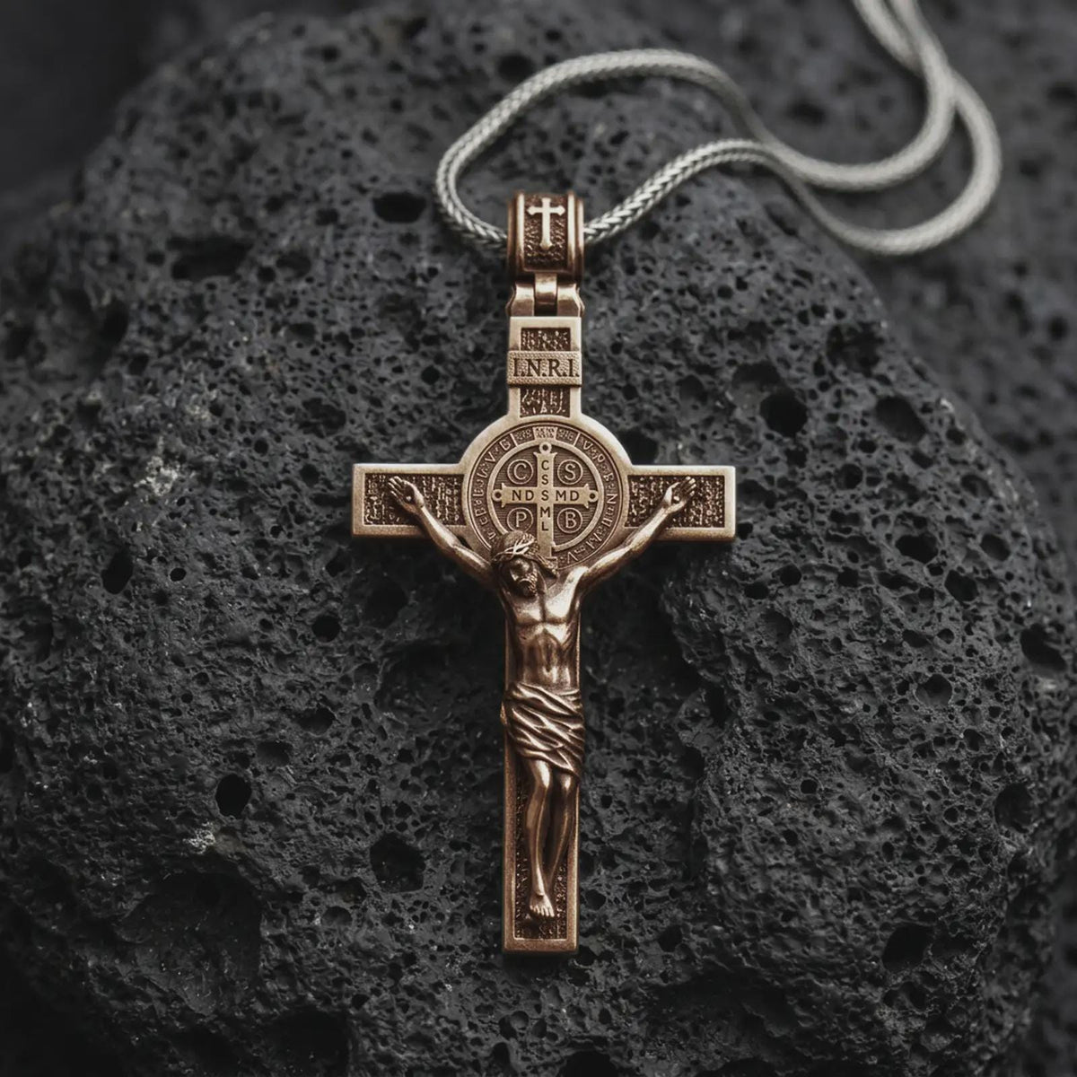An bronze sterling silver Saint Benedict crucifix rests on dark volcanic rock, featuring the sculpted Jesus figure, INRI inscription, central Benedict medal, and textured Catholic cross details.