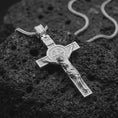 Load image into Gallery viewer, The polished silver Saint Benedict crucifix is displayed on black volcanic rock, highlighting the raised Jesus figure, detailed central medal, INRI plaque, and geometric cross frame.
