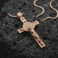 Load image into Gallery viewer, The rose gold plated Saint Benedict crucifix is arranged on dark volcanic rock, emphasizing the sculpted Christ figure, Benedict medal centerpiece, raised borders, and Christian religious details.
