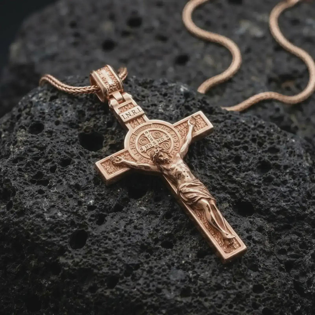 The rose gold plated Saint Benedict crucifix is arranged on dark volcanic rock, emphasizing the sculpted Christ figure, Benedict medal centerpiece, raised borders, and Christian religious details.