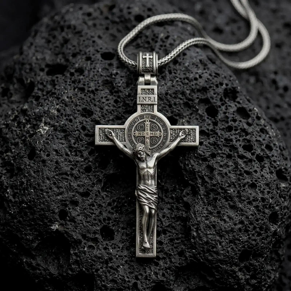 An oxidized sterling silver Saint Benedict crucifix rests on dark volcanic rock, featuring the sculpted Jesus figure, INRI inscription, central Benedict medal, and textured Catholic cross details.