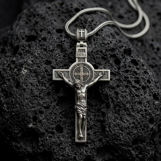 An oxidized sterling silver Saint Benedict crucifix rests on dark volcanic rock, featuring the sculpted Jesus figure, INRI inscription, central Benedict medal, and textured Catholic cross details.