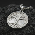 Load image into Gallery viewer, The polished sterling silver Yggdrasil necklace is displayed on textured black volcanic rock, highlighting the sculpted tree trunk, spreading branches, delicate foliage, intertwined roots, and smooth circular border.

