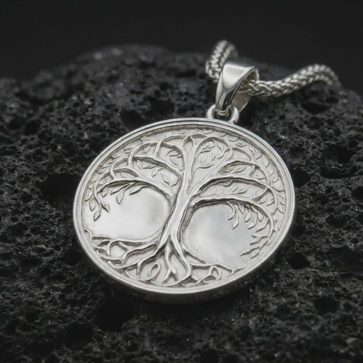 The polished sterling silver Yggdrasil necklace is displayed on textured black volcanic rock, highlighting the sculpted tree trunk, spreading branches, delicate foliage, intertwined roots, and smooth circular border.