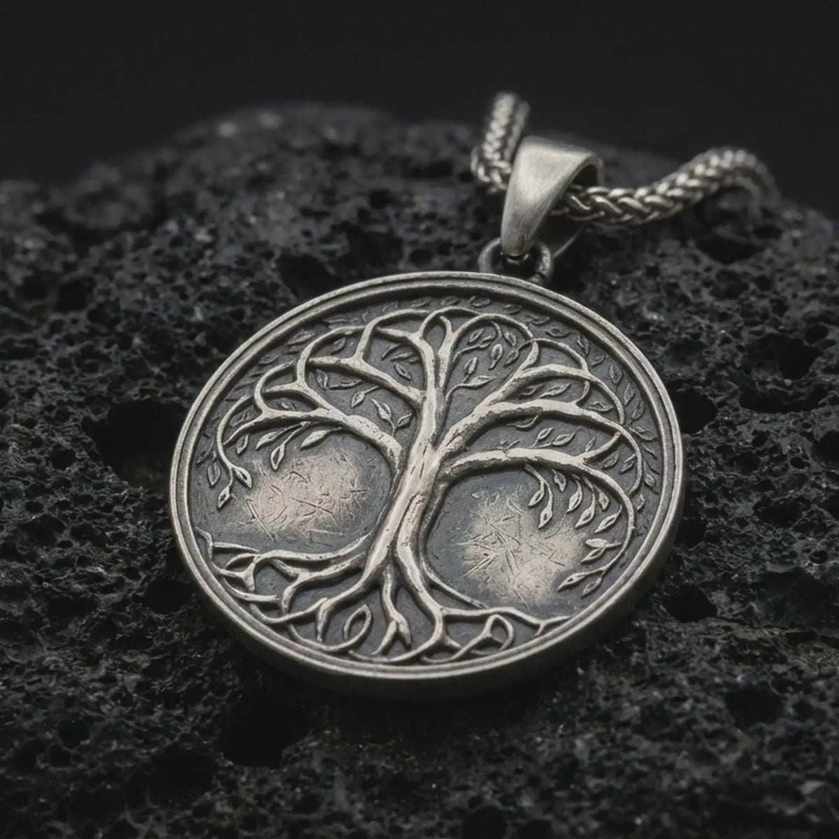 An oxidized sterling silver Tree of Life pendant rests on dark volcanic rock, featuring raised Yggdrasil branches, delicate leaves, intertwined roots, and a circular medallion with dark recessed details.
