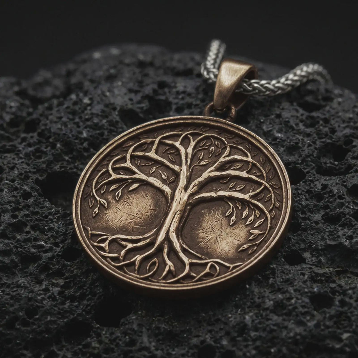 A bronze sterling silver Tree of Life pendant rests on dark volcanic rock, featuring raised Yggdrasil branches, delicate leaves, intertwined roots, and a circular medallion with dark recessed details.