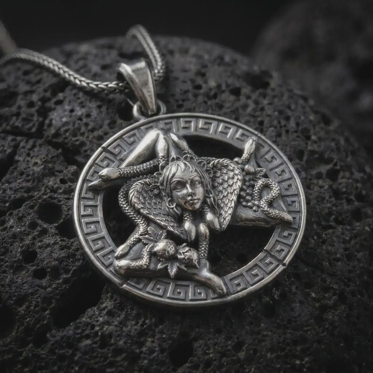 Oxidized sterling silver Trinacria pendant on black volcanic rock, featuring Medusa, three bent legs, and a detailed Greek key border inspired by the traditional symbol of Sicily.