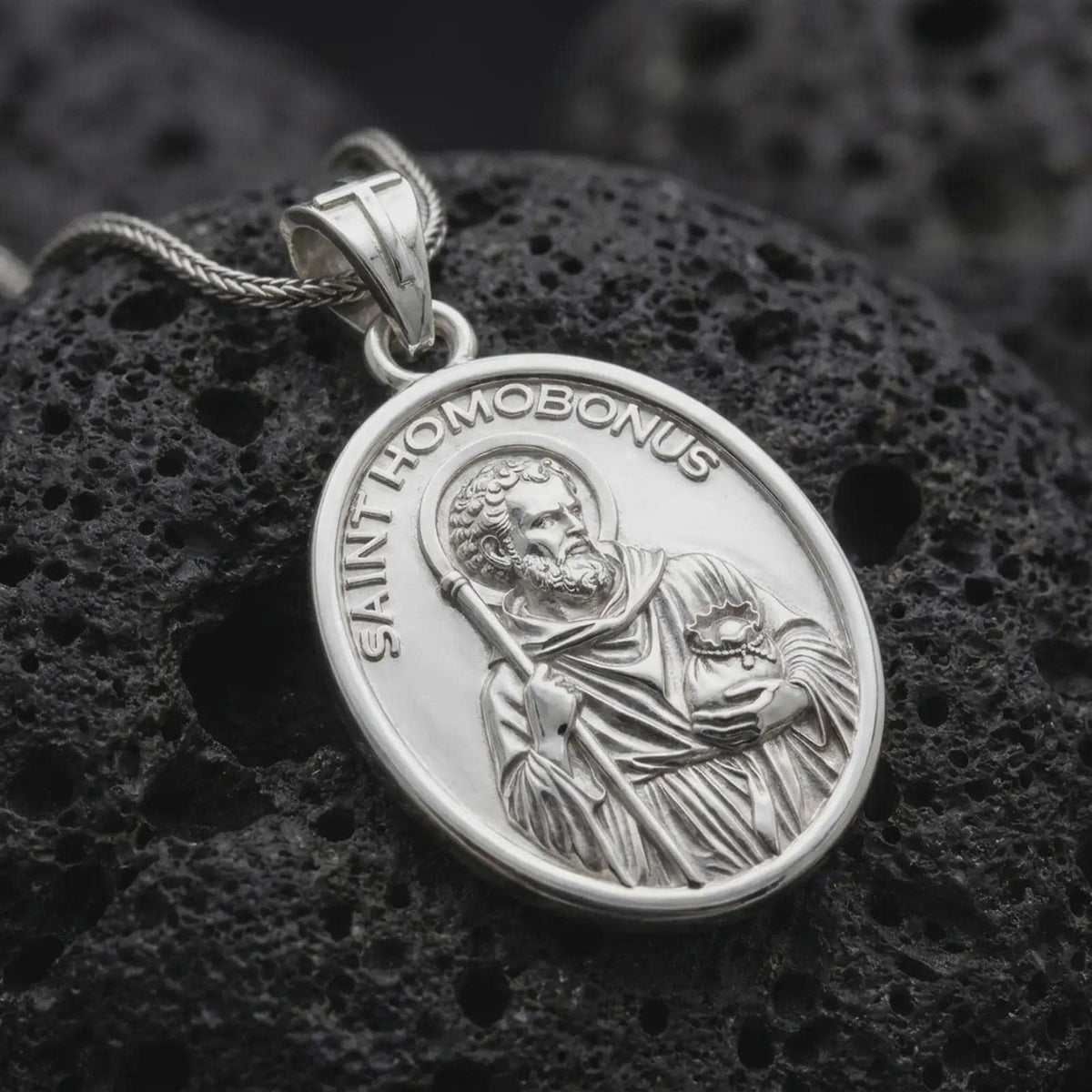Polished sterling silver Saint Homobonus pendant displayed on dark volcanic stone, highlighting the saint portrait, halo, staff, religious details, and oval medal frame.