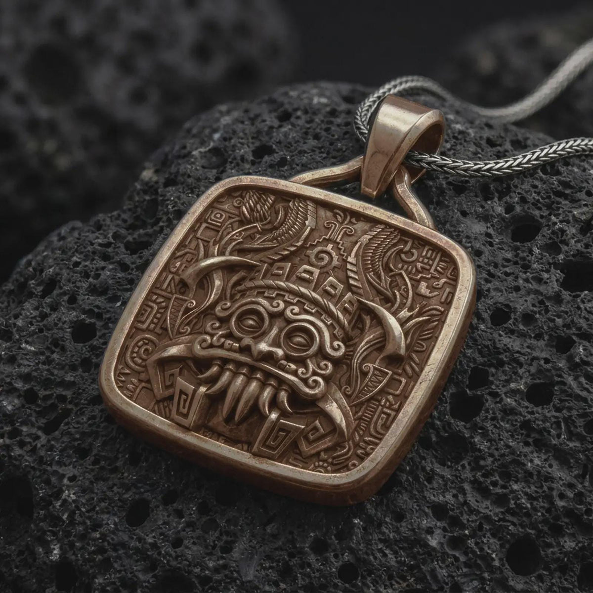 Bronze Tlaloc pendant on black volcanic rock, featuring ringed eyes, prominent fangs, curled facial details, and intricate Aztec geometric patterns.