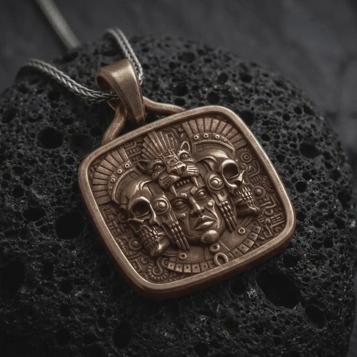 Bronze Aztec jaguar pendant on black volcanic rock, featuring a ceremonial human face, feline headdress, flanking skulls, feathers, and intricate Mesoamerican relief details.