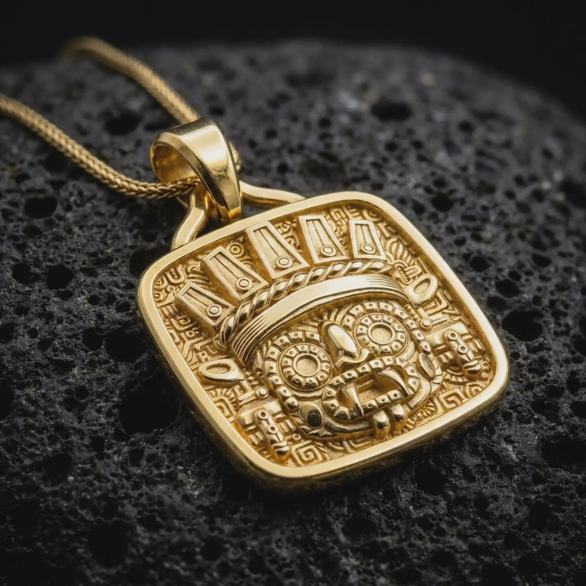 Gold plated Tlaloc pendant resting on rugged black volcanic rock, featuring the Aztec rain deity with goggle eyes, fangs, ceremonial ornamentation, and intricate Mesoamerican motifs.