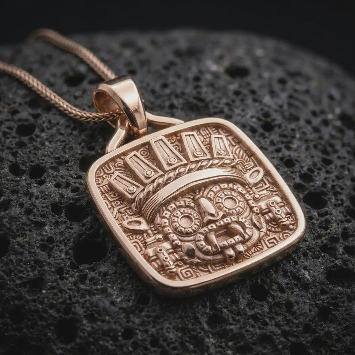 Rose gold plated Tlaloc necklace displayed on textured volcanic stone, showcasing the sculpted rain god face, detailed headdress, circular eyes, and geometric Aztec relief.