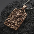 Load image into Gallery viewer, Bronze horned demon skull pendant on black volcanic rock, featuring large curled ram horns, skeletal hands, flames, and a smaller skull in detailed gothic relief.
