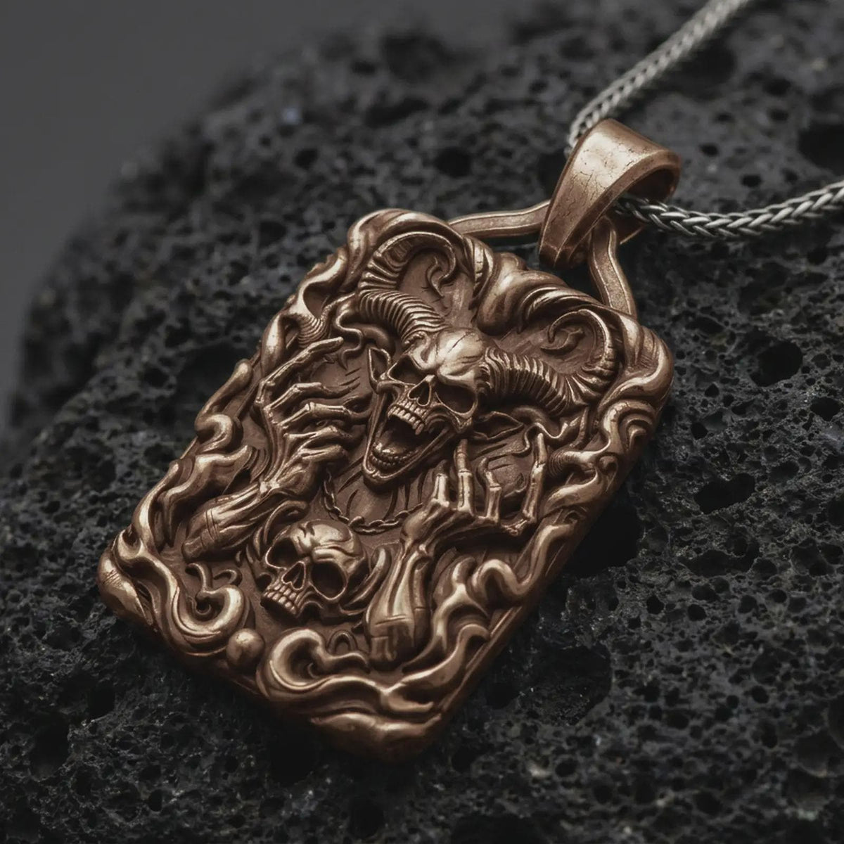 Bronze horned demon skull pendant on black volcanic rock, featuring large curled ram horns, skeletal hands, flames, and a smaller skull in detailed gothic relief.