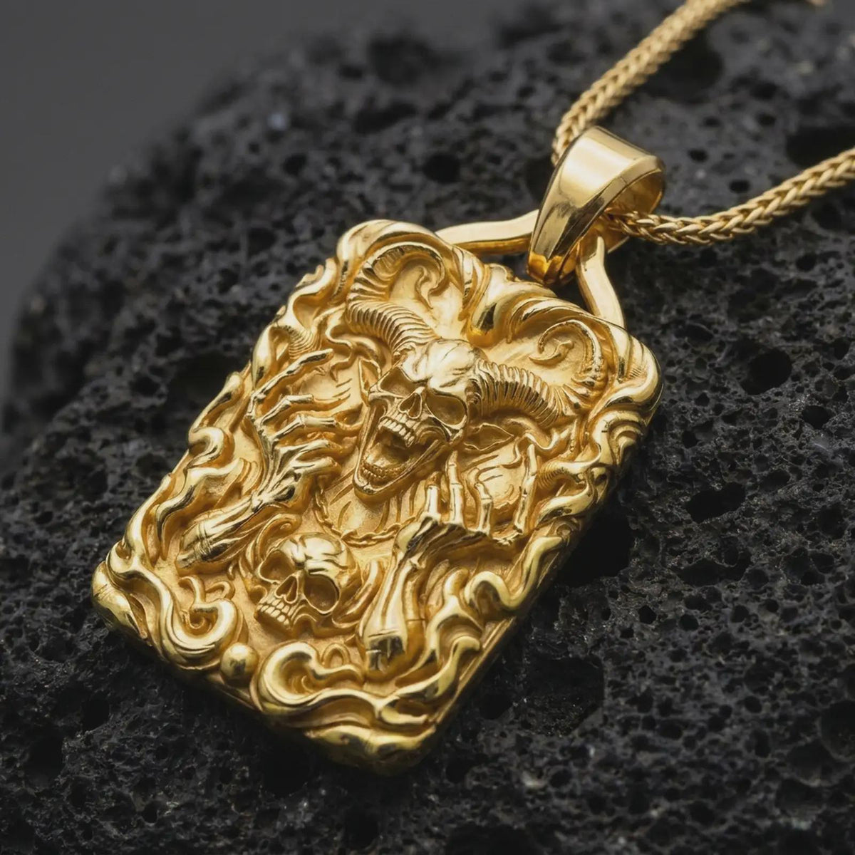 Gold plated horned skull pendant on black volcanic rock, featuring curled ram horns, an open-mouthed demon skull, skeletal hands, flames, and detailed gothic ornamentation.