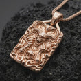 Load image into Gallery viewer, Rose gold plated demon skull necklace displayed on textured volcanic stone, showcasing the ram-horn skull, skeletal imagery, flames, and layered dark fantasy relief.
