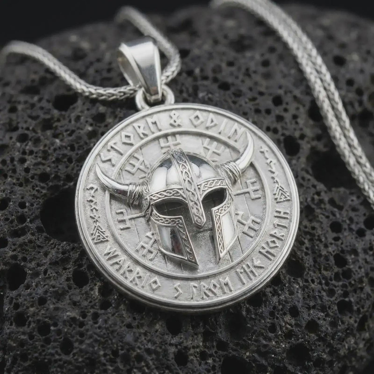Polished sterling silver Odin necklace displayed on dark volcanic stone, highlighting the horned Viking helmet, engraved symbols, circular medallion frame, and warrior design.