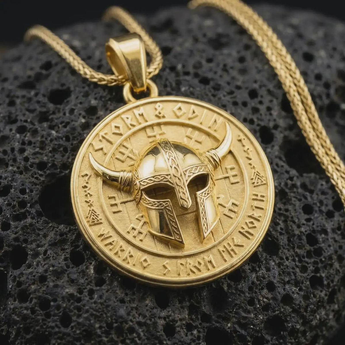 Gold plated Viking helmet pendant resting on black volcanic rock, featuring a detailed horned helmet, Norse-inspired runes, Storm and Odin inscription, and round medallion design.