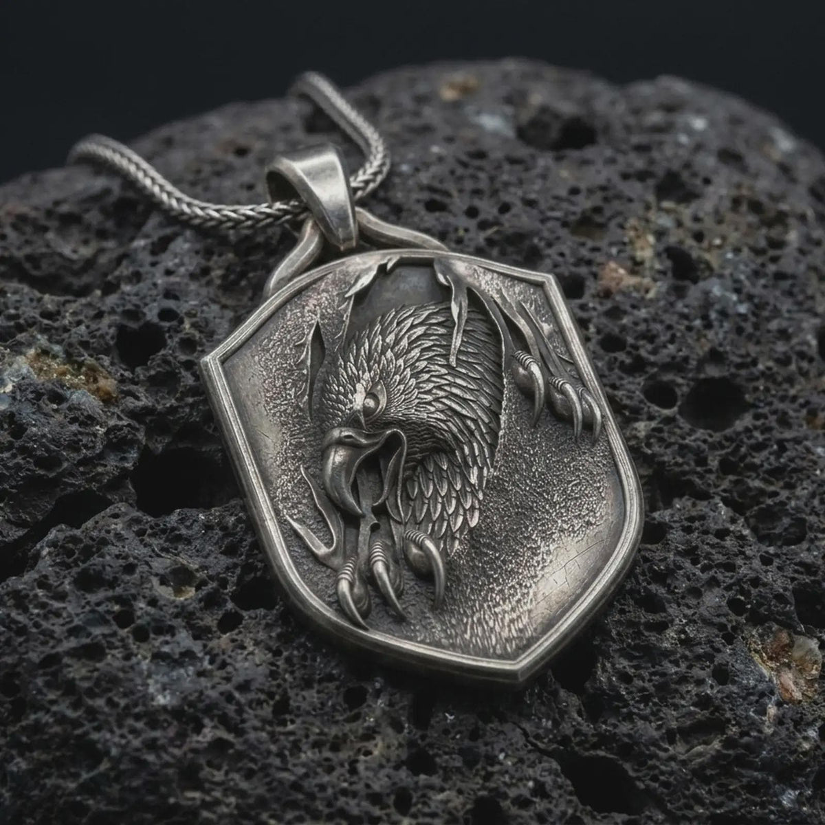 Oxidized sterling silver eagle shield pendant on black volcanic rock, featuring a detailed eagle head, layered feathers, sharp talons, and textured heraldic shield.