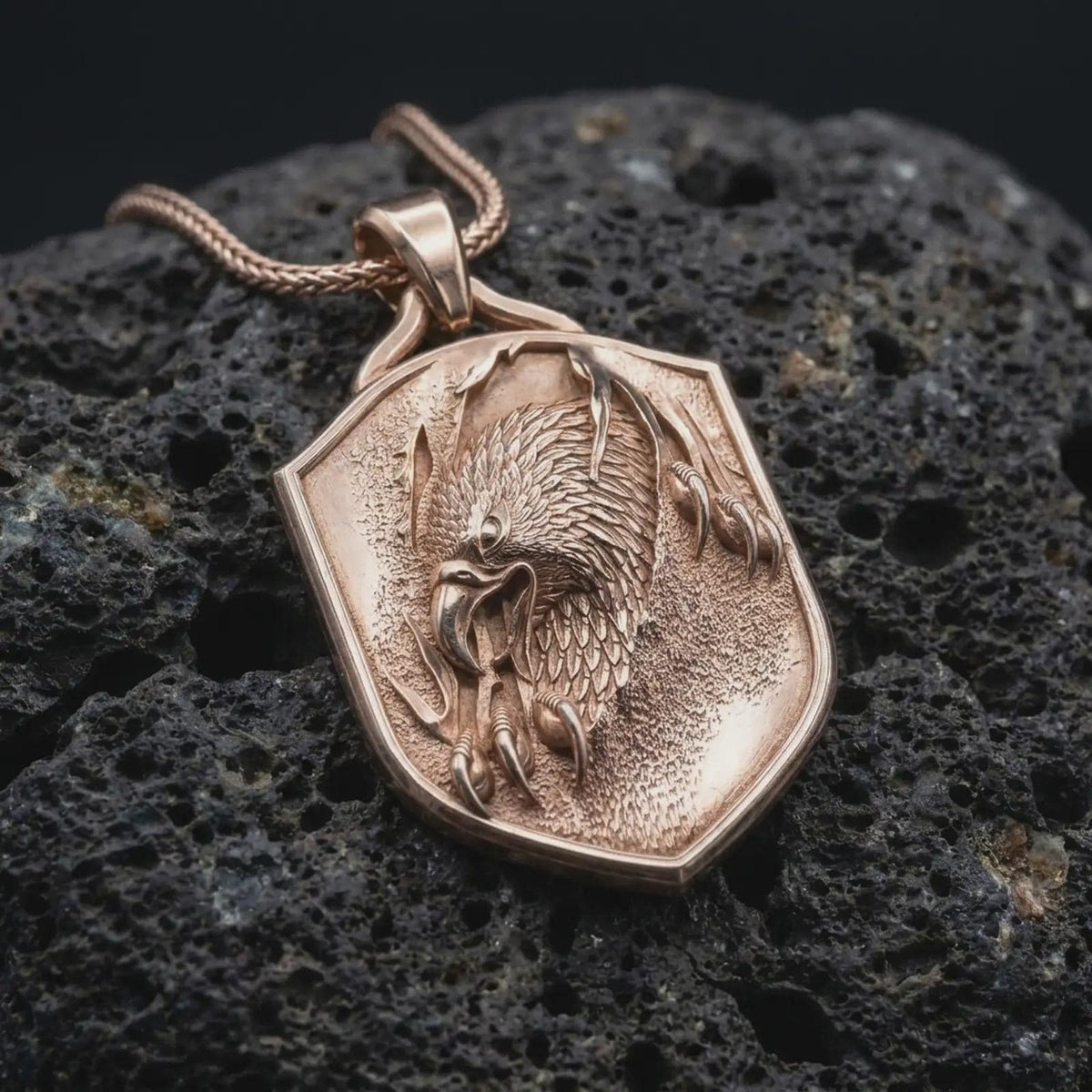 Rose gold plated eagle necklace displayed on textured volcanic stone, showcasing the sculpted eagle head, layered feathers, talon details, and distinctive shield pendant shape.