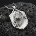 Load image into Gallery viewer, Polished sterling silver eagle necklace displayed on dark volcanic stone, highlighting the sculpted bird of prey head, feather details, curved talons, and shield-shaped frame.
