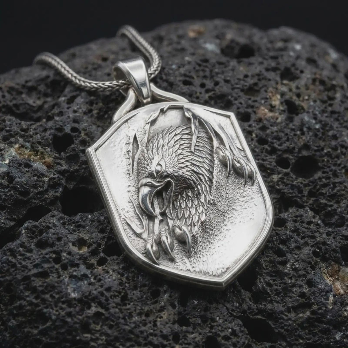 Polished sterling silver eagle necklace displayed on dark volcanic stone, highlighting the sculpted bird of prey head, feather details, curved talons, and shield-shaped frame.