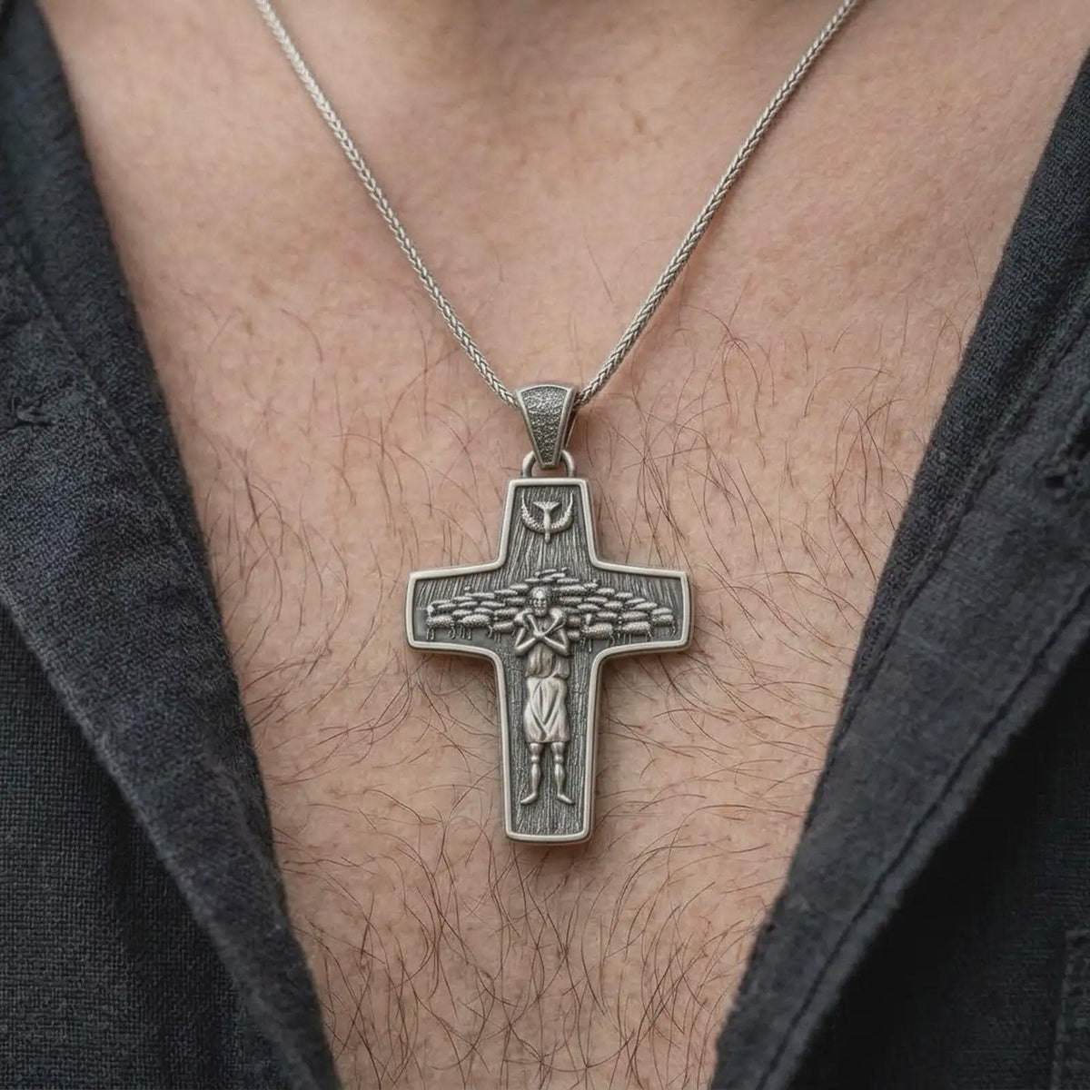 An oxidized sterling silver Pope Francis cross pendant is worn on the chest, displaying the Good Shepherd carrying a sheep, surrounding flock, dove motif, and substantial Christian cross profile in a close-up lifestyle photograph.