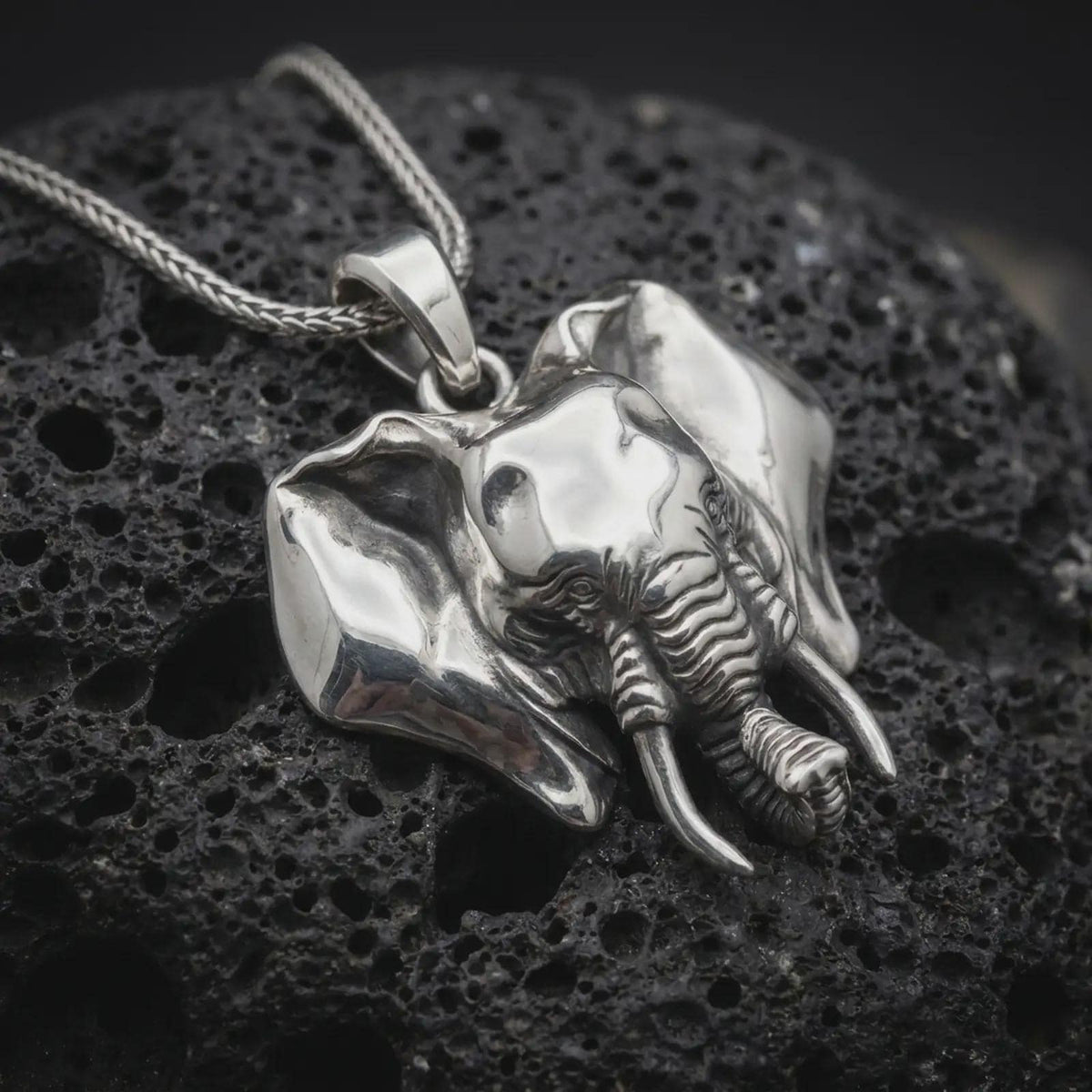 Polished sterling silver elephant necklace displayed on dark volcanic stone, highlighting the realistic elephant head, broad ears, curved tusks, textured skin details, and sculpted trunk.