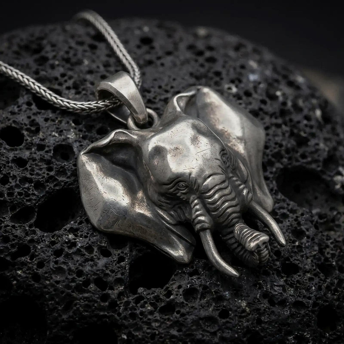 Oxidized sterling silver elephant head pendant on black volcanic rock, featuring large sculpted ears, detailed tusks, expressive eyes, and a dimensional curled trunk.