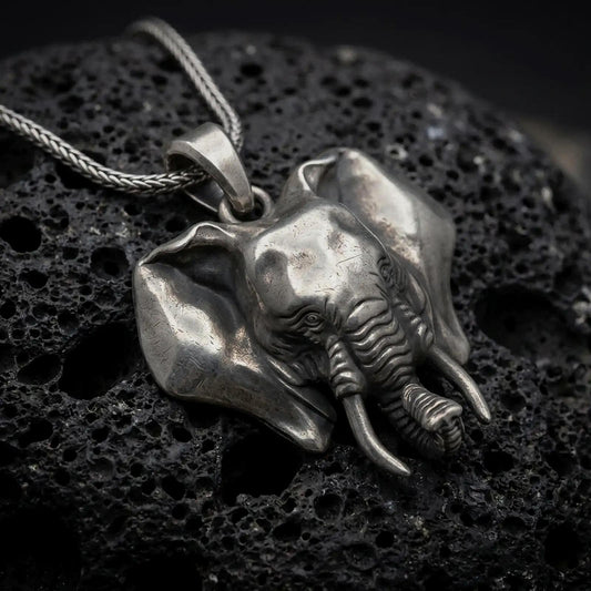 Oxidized sterling silver elephant head pendant on black volcanic rock, featuring large sculpted ears, detailed tusks, expressive eyes, and a dimensional curled trunk.