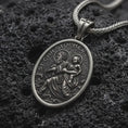 Load image into Gallery viewer, An oxidized sterling silver St. Christopher pendant rests on dark volcanic rock, featuring Saint Christopher carrying the Christ Child across flowing water with halo details and raised Catholic religious relief in a macro studio close-up.
