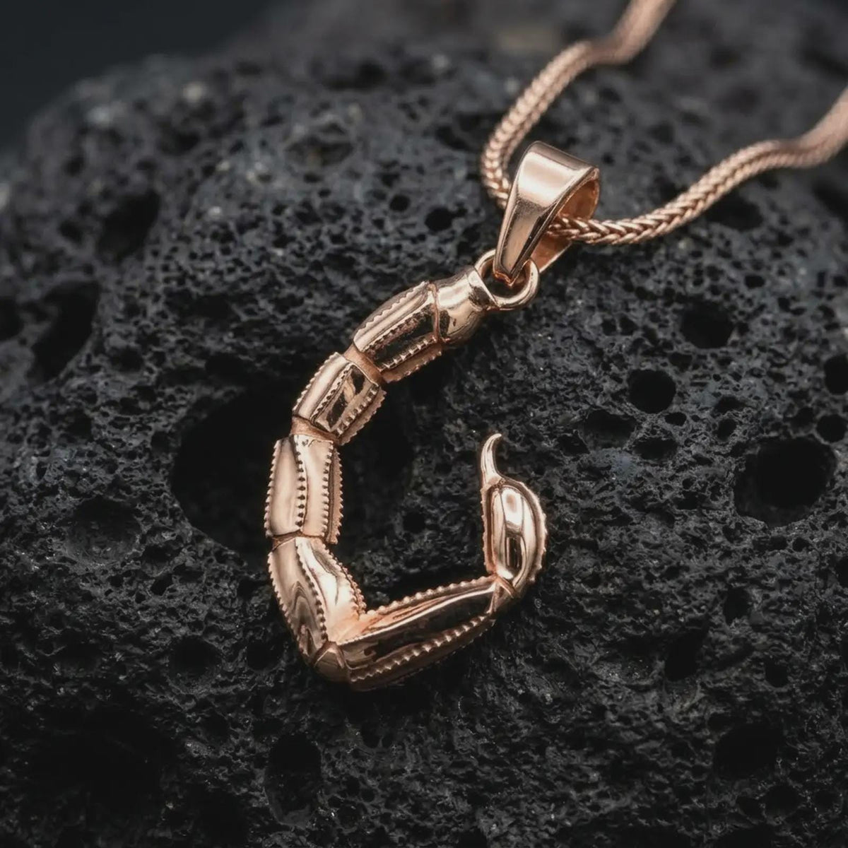 The rose gold plated Scorpio tail necklace is arranged on black volcanic rock, emphasizing the segmented sculptural form, pointed stinger, and distinctive zodiac-inspired statement pendant design in a refined macro image.
