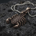 Load image into Gallery viewer, A bronze sterling silver scorpion pendant rests on dark volcanic rock, featuring detailed claws, segmented legs, textured body armor, curved tail, and pointed stinger in a dramatic macro studio close-up.
