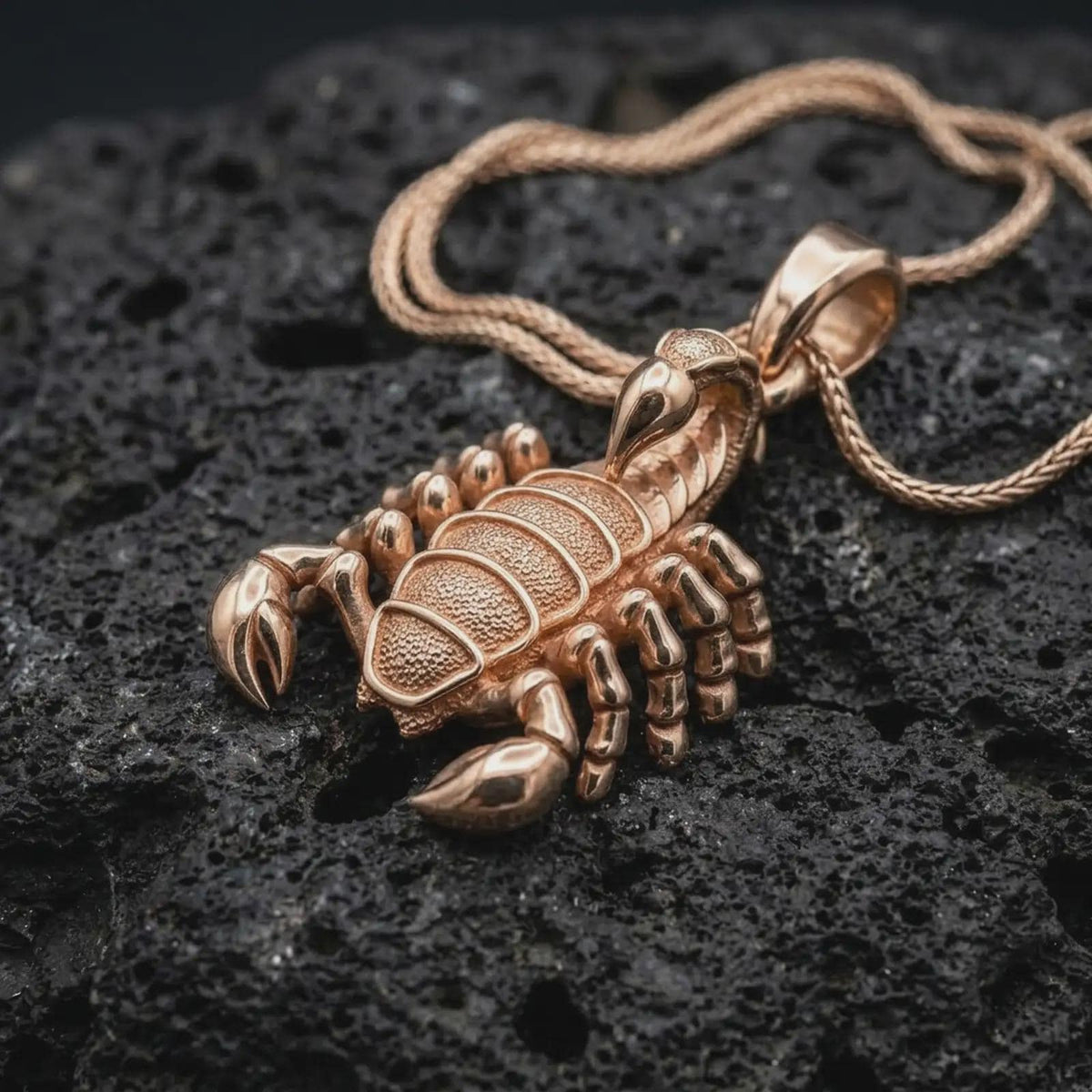The rose gold plated scorpion pendant is arranged on black volcanic rock, emphasizing its three-dimensional animal form, sculpted claws, articulated legs, textured abdomen, and pointed stinger.