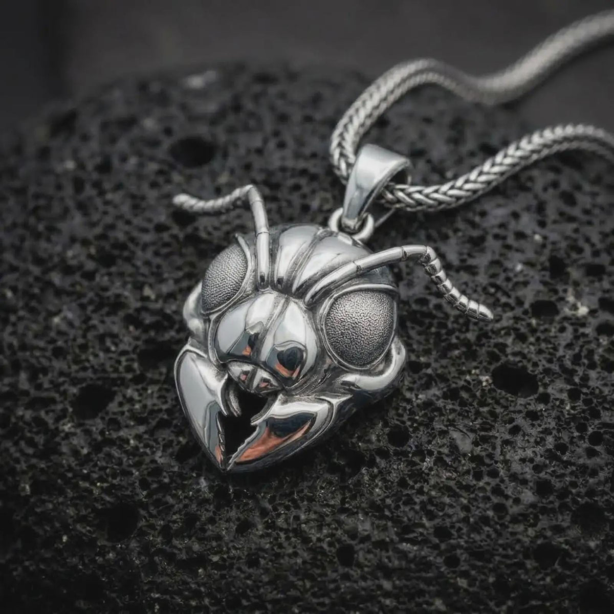 The polished sterling silver insect pendant is displayed on black volcanic rock, highlighting the reflective ant head, textured eyes, articulated antennae, and dimensional pincer-like mandibles in a premium macro photograph.