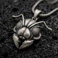 Load image into Gallery viewer, An oxidized sterling silver ant head pendant rests on dark volcanic rock, featuring large textured compound eyes, segmented antennae, sculpted facial armor, and prominent mandibles in a dramatic macro close-up.

