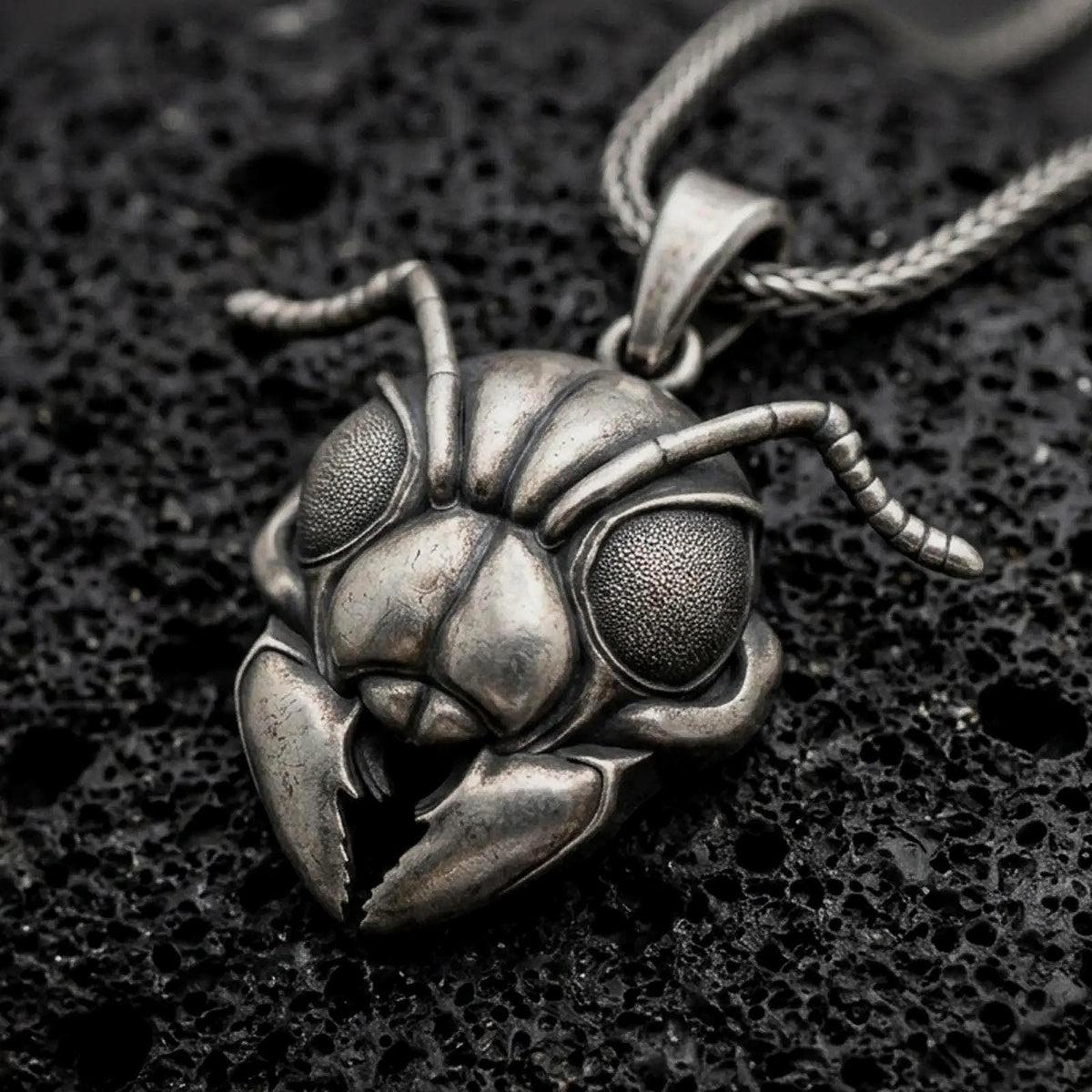 An oxidized sterling silver ant head pendant rests on dark volcanic rock, featuring large textured compound eyes, segmented antennae, sculpted facial armor, and prominent mandibles in a dramatic macro close-up.