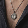 Load image into Gallery viewer, An oxidized sterling silver Yggdrasil pendant is worn on the chest, displaying the circular Tree of Life medallion, dark recessed background, intricate branches, leaves, and intertwined roots in a close-up lifestyle photograph.
