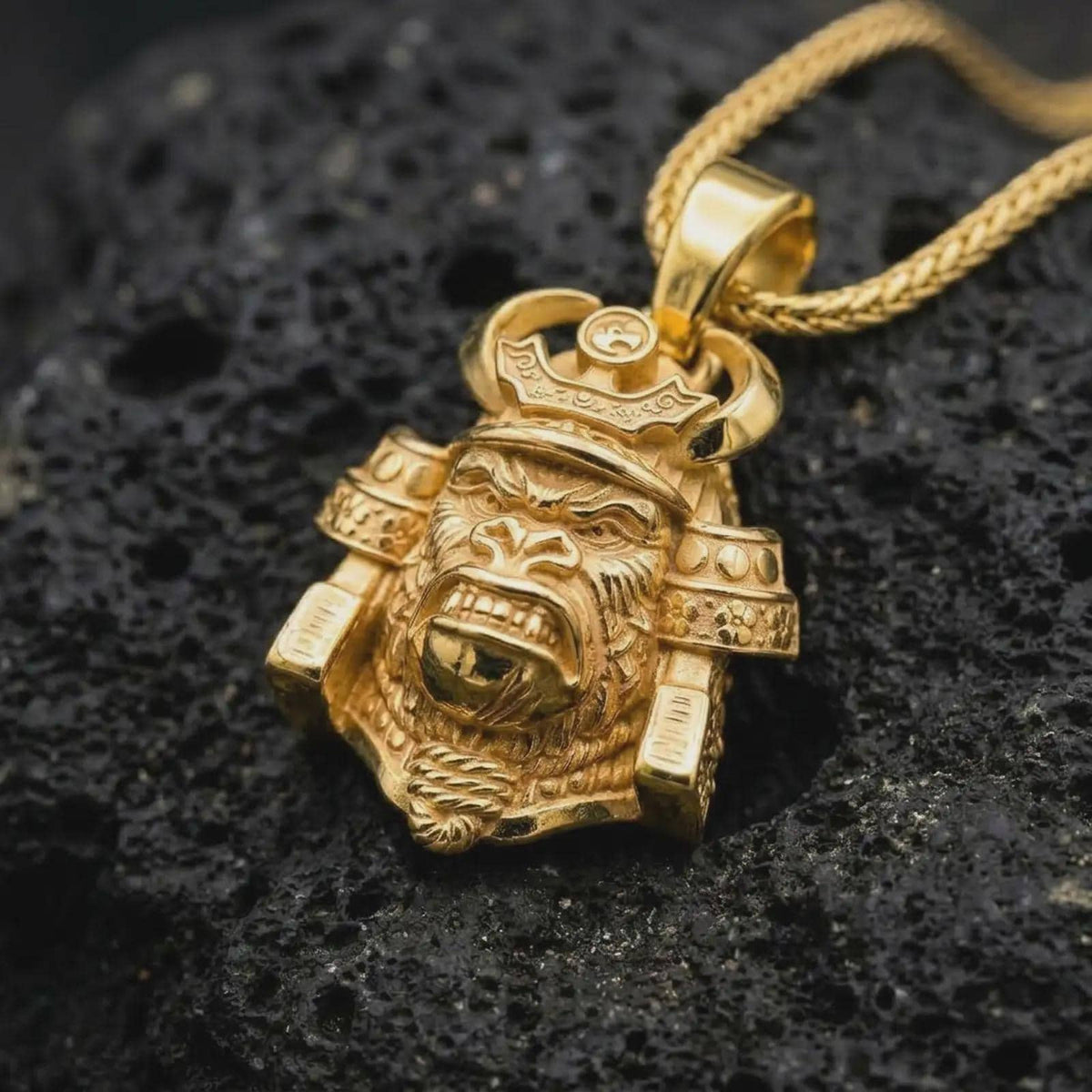 A gold plated King Gorilla Samurai necklace rests on rugged black volcanic rock, showcasing the sculpted gorilla head, curved samurai helmet horns, decorative crown, and detailed warrior armor.