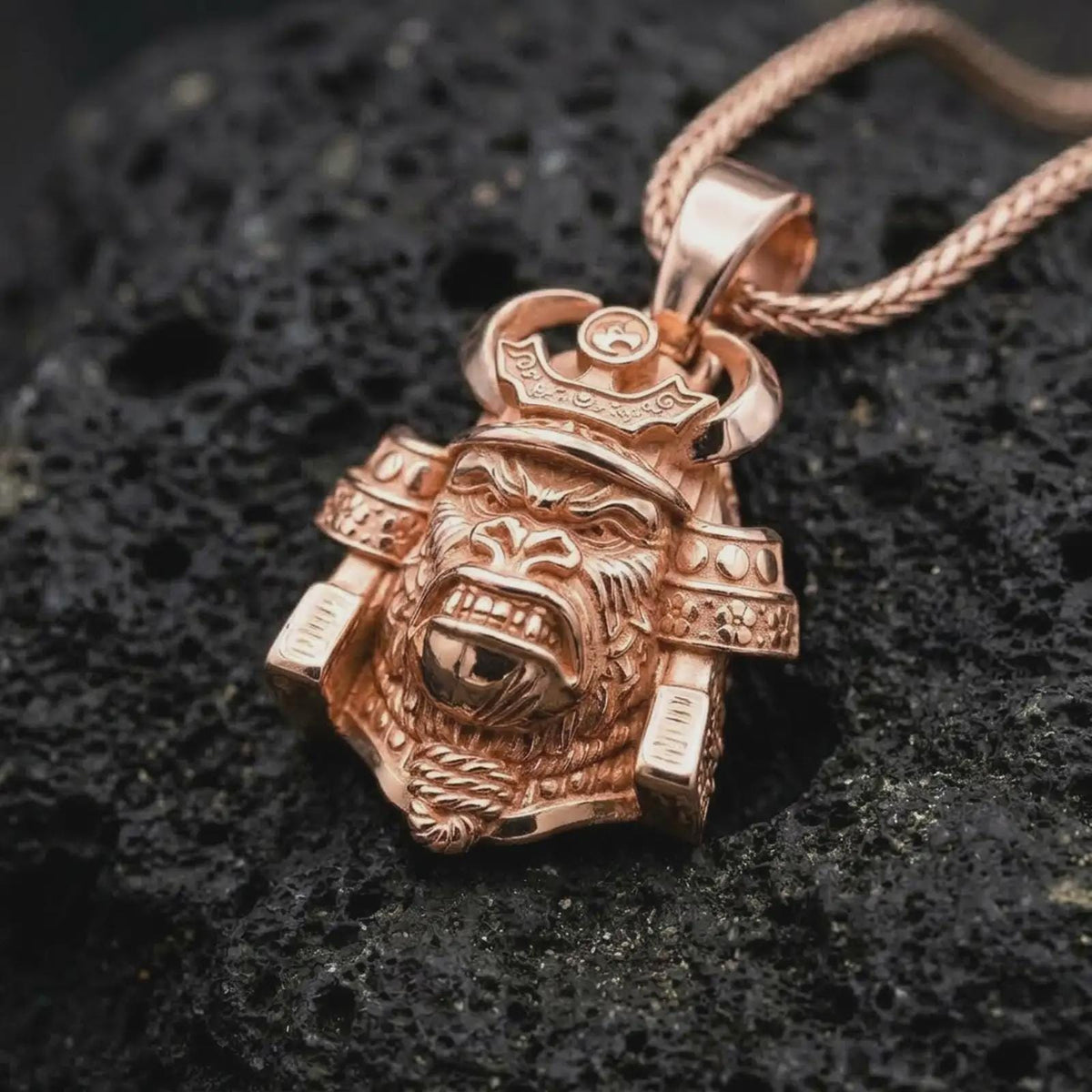 A rose gold plated gorilla samurai pendant displayed on textured volcanic rock, featuring an aggressive gorilla expression, ornamental helmet details, raised metal textures, and bold warrior-inspired design.